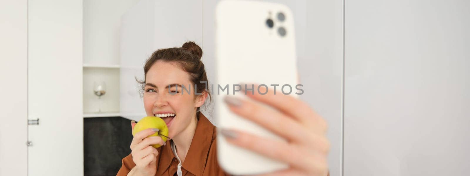 Portrait of happy, smiling young woman records herself, takes selfie while eating an apple in the kitchen, using smartphone app, makes photos with mobile phone.