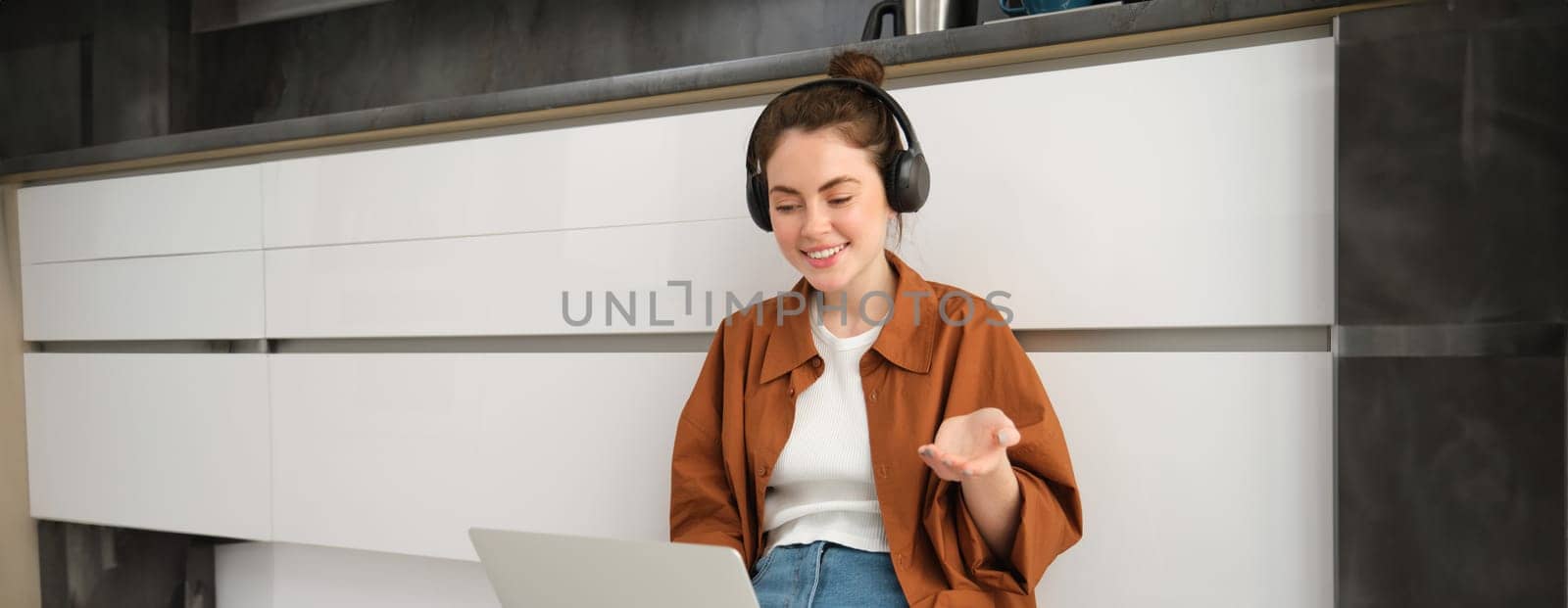 Portrait of young smiling woman explains something over the video call, sits in headphones with laptop, works from home by Benzoix
