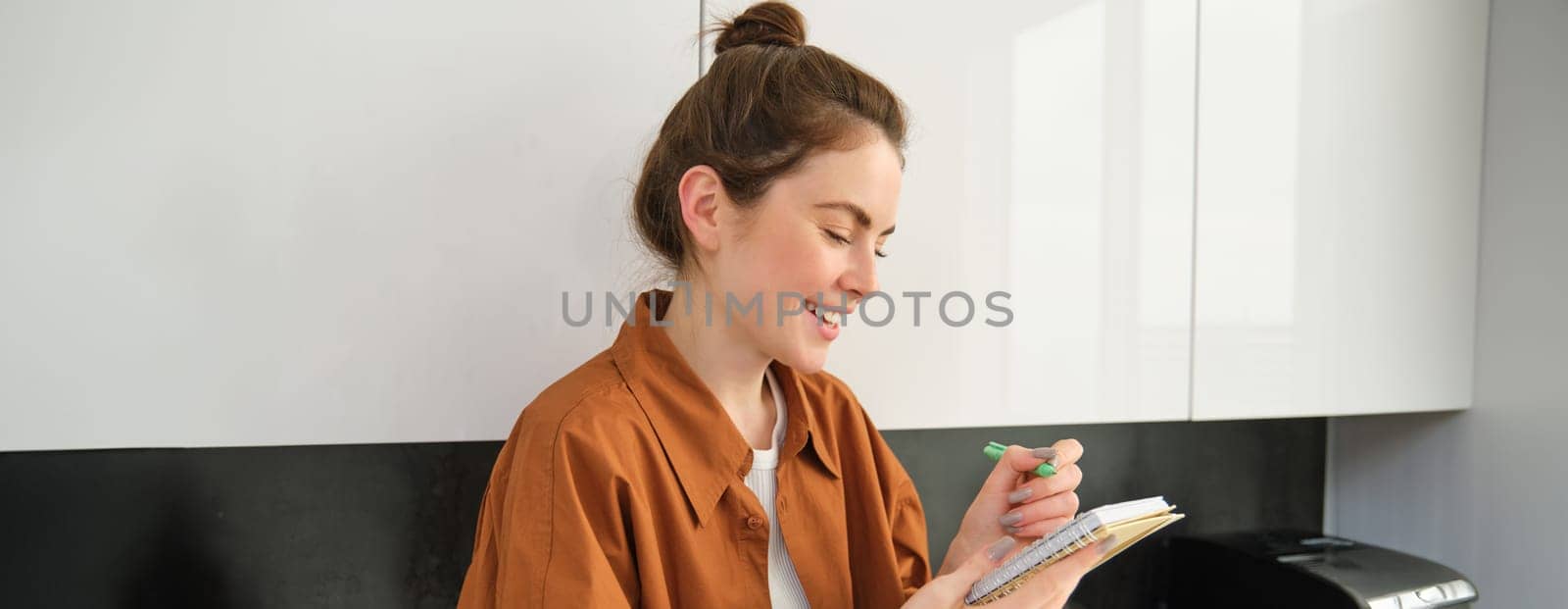 Portrait of happy cute woman at home, writing in notebook, makes notes in her diary, has a planner in her hands, sits in kitchen.
