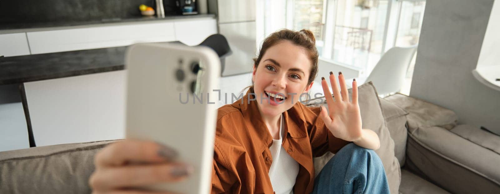Friendly smiling young woman, sitting on couch and waving hand at smartphone camera, video chats, talking on mobile phone, connects to online conversation on application.
