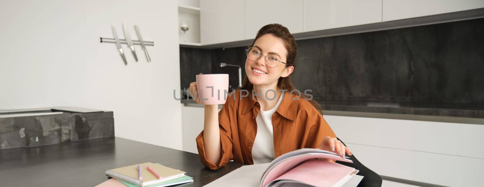 Portrait of beautiful student, woman in glasses reading and drinking coffee at home, studying and revising for lesson, doing homework.