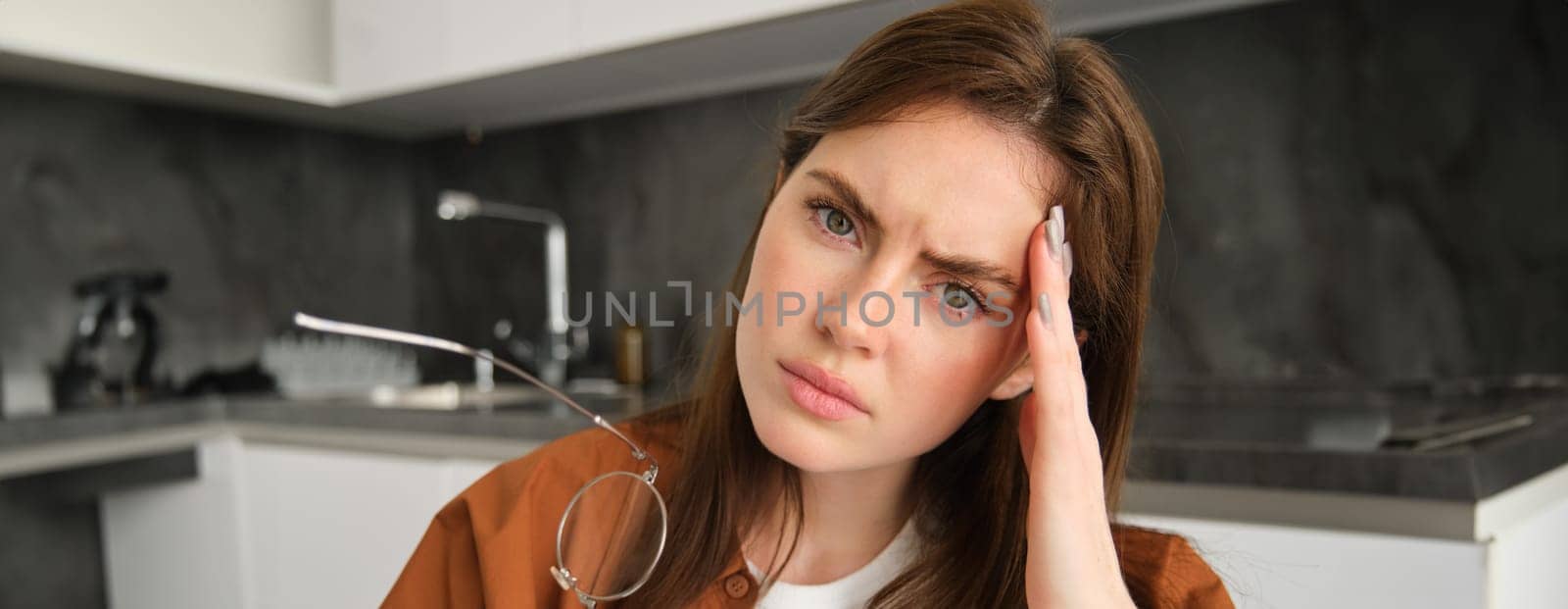 Close up portrait of woman frowning, touching head from painful feeling, has headache or migraine, troubled with something, sitting in kitchen.