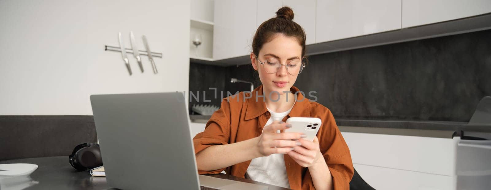 Portrait of young female freelancer, woman working from home with laptop, girl programmer sitting with computer in kitchen, answer message on smartphone.