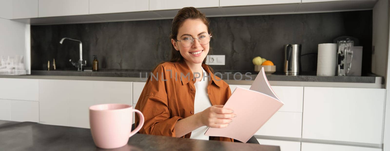 Portrait of smiling girl, student in glasses in kitchen, holding folder, doing homework, revising for test, reading document.