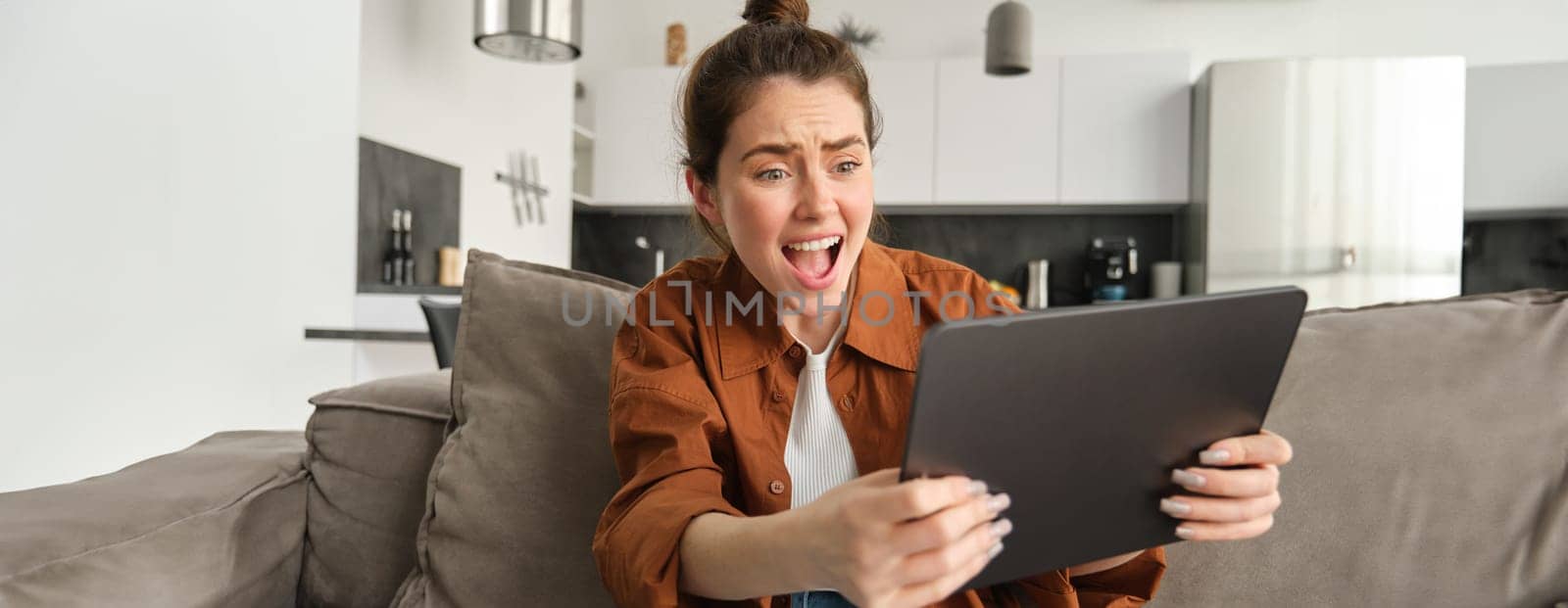 Portrait of woman looking at digital tablet with worried, stressed and upset face, losing in video game, sitting at home on couch.