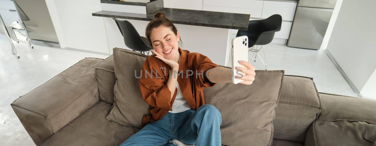 Portrait of beautiful smiling woman, takes selfie at home, posing on sofa, holds smartphone with extended hand.