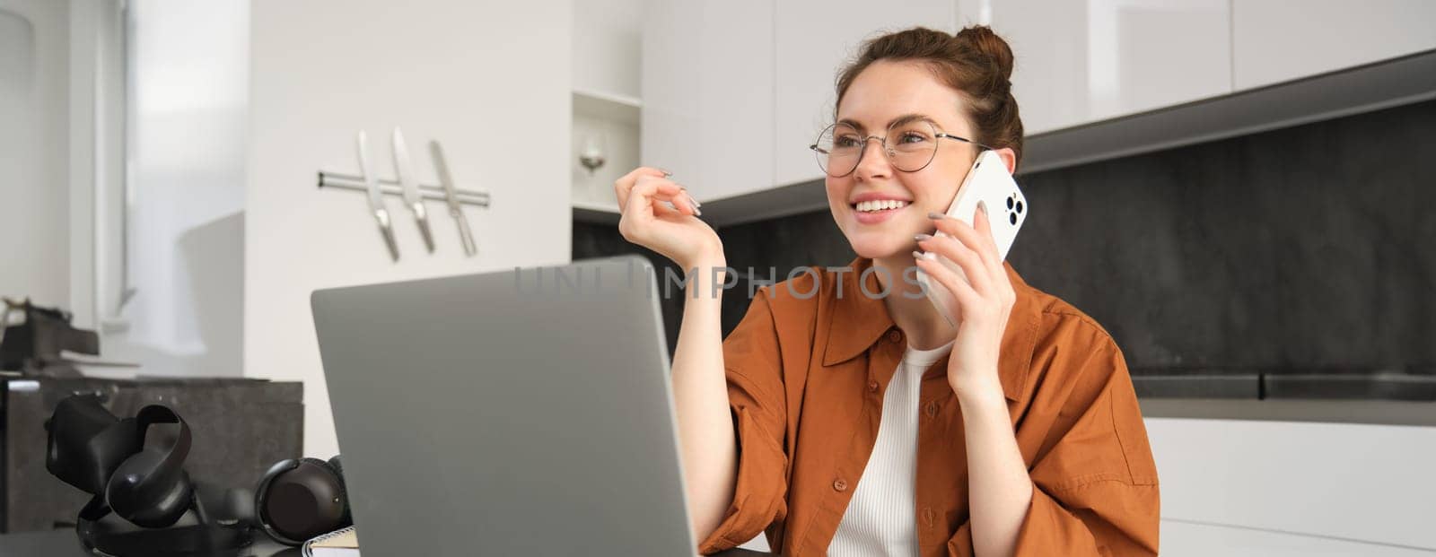 Portrait of young self-employed woman, entrepreneur working from home, freelancer calling client. Girl making an order, talking to someone on phone, sitting in kitchen with laptop by Benzoix