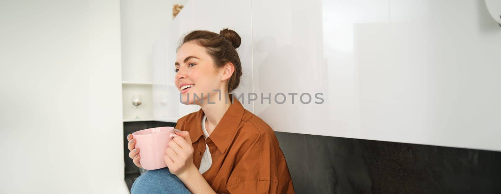 Cosy moments. Happy young woman sits in kitchen and drinks coffee, has tender smile on her face.