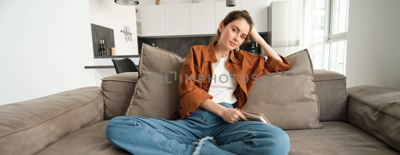 Lifestyle concept. Young smiling brunette woman, sitting at home on sofa, resting on couch, looking at camera with relaxed expression.