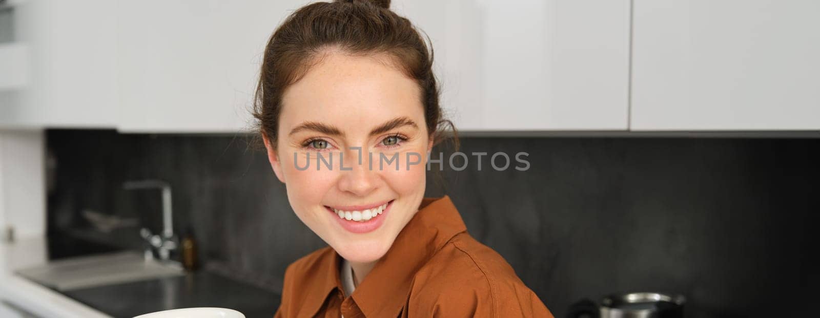 Portrait of smiling gorgeous woman enjoys cuppa at home. Cute girl with cup of coffee, drinking cappuccino in kitchen.