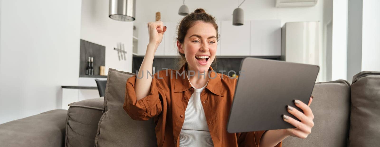 Excited cute woman winning, celebrating, looking at digital tablet and rejoicing, triumphing, reading great news, sitting on couch at home by Benzoix