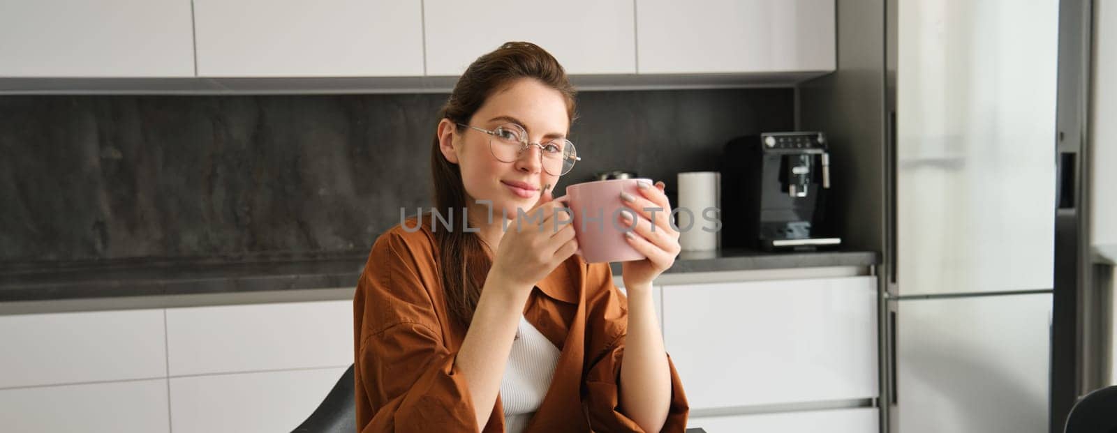 Portrait of beautiful brunette woman, sitting in kitchen, taking a break for cup of coffee. Girl drinks tea at home.
