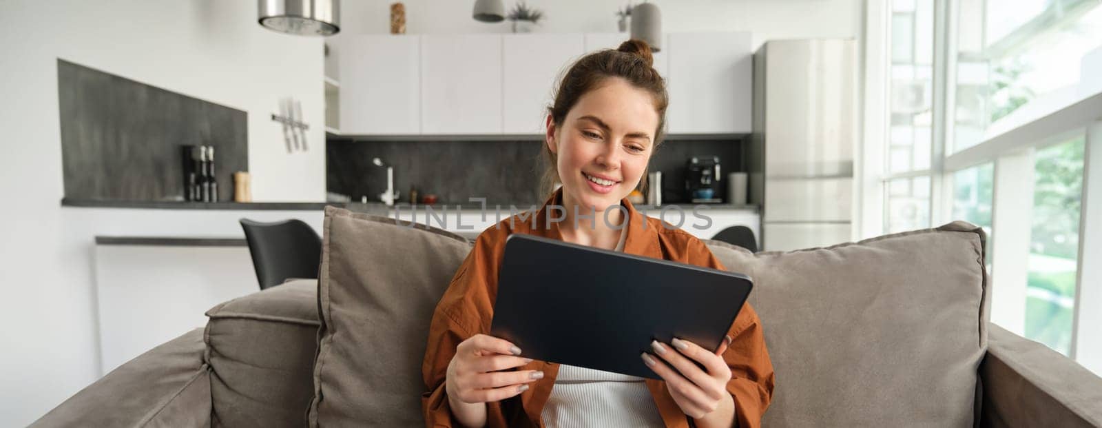 Portrait of young brunette woman reading on digital tablet, watching tv series on her application, sitting on couch in living room by Benzoix