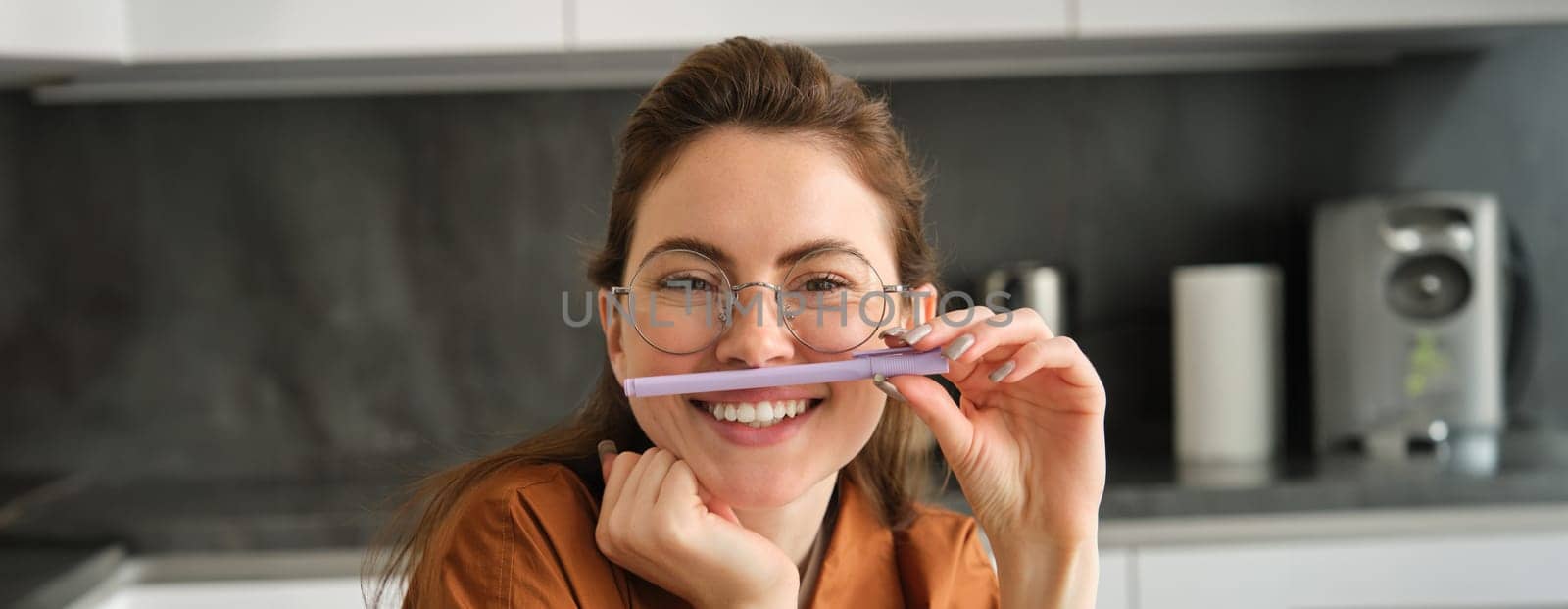 Close up portrait of funny, happy young woman, student sitting in kitchen, learning on remote and playing with pen.