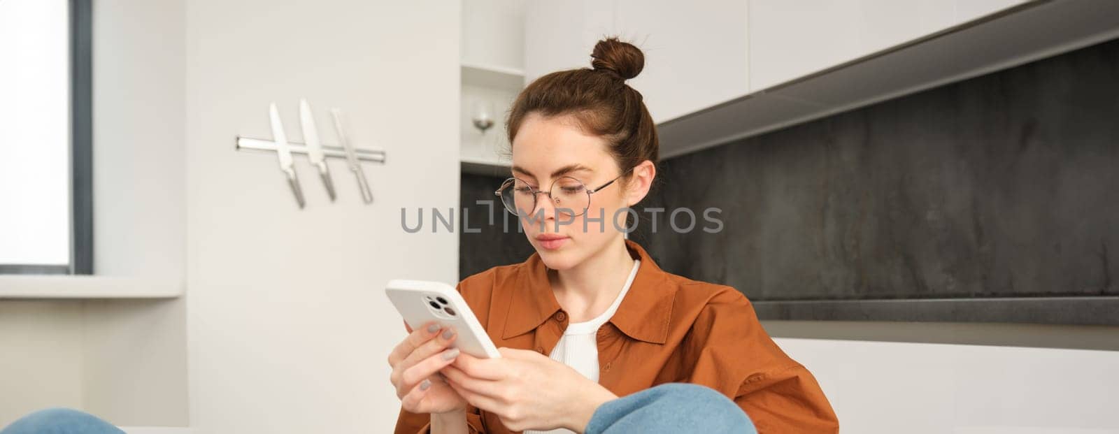 Portrait of young brunette woman in glasses, sitting at home with mobile phone, using her smartphone and smiling, reading message.