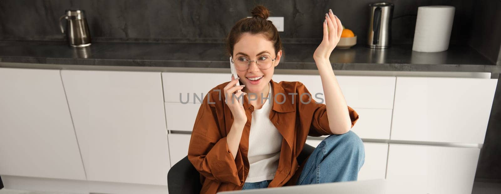 Excited young woman talking on mobile phone in front of laptop, sitting in kitchen with happy face expression, having a conversation by Benzoix
