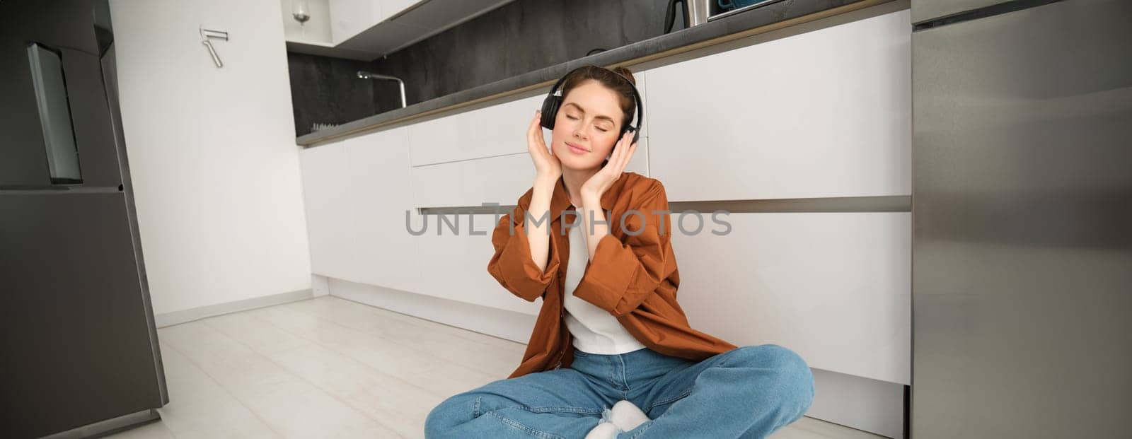 Portrait of young beautiful woman in wireless headphones, sitting on kitchen floor, vibing with favourite music in earphones.