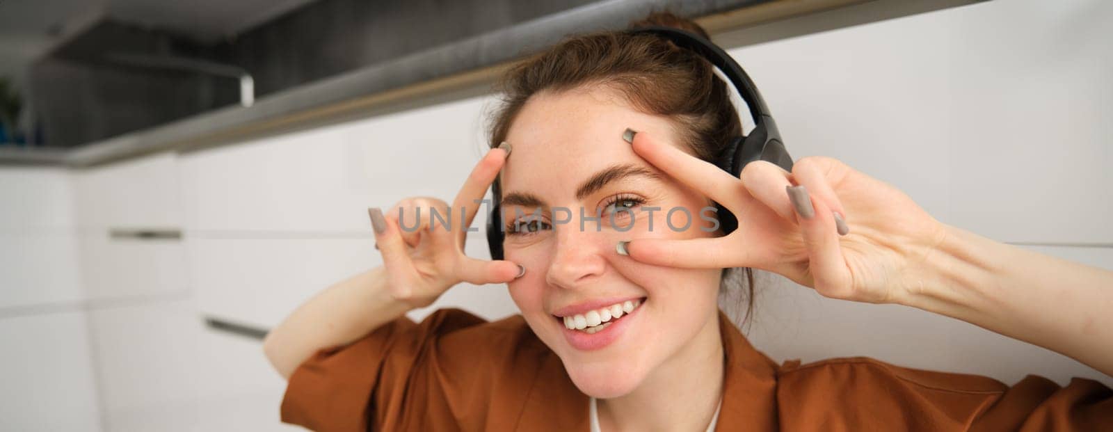 Close up of carefree young woman, laughing and smiling, showing peace, v-sign gesture, listening to music in wireless headphones.