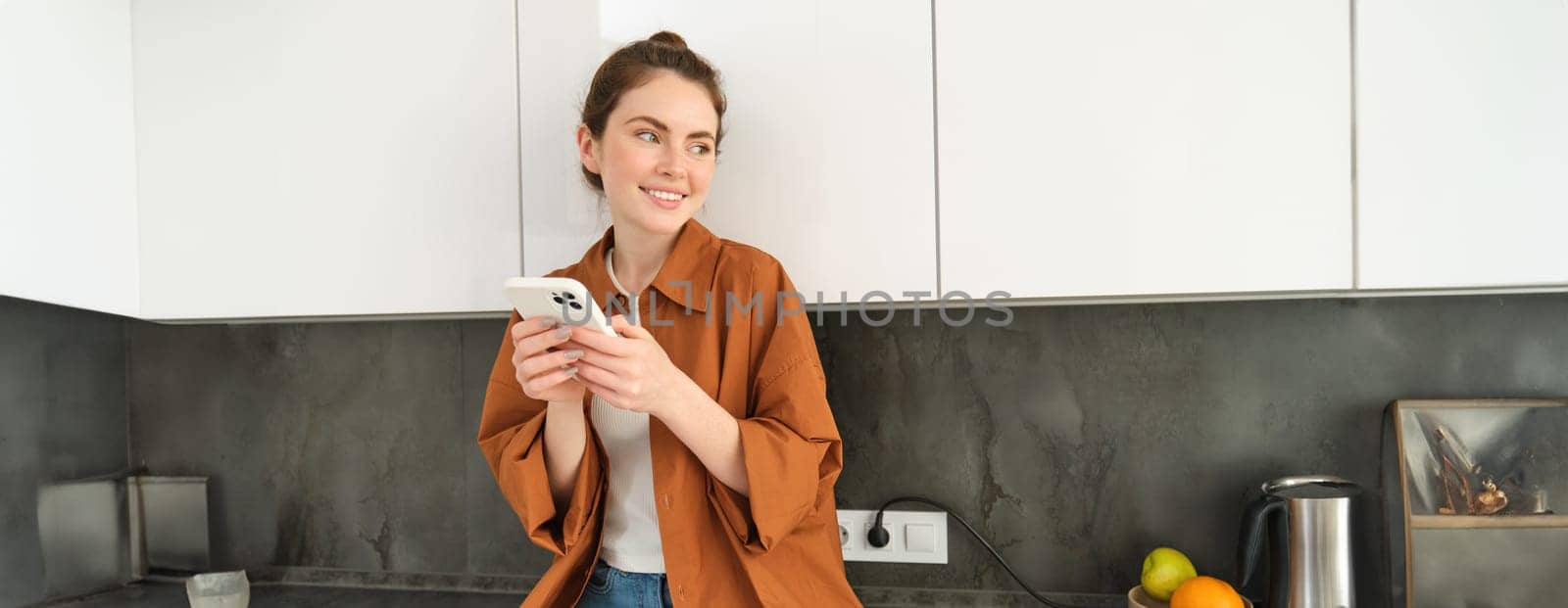 Modern girl at home, sits on kitchen counter with smartphone and smiling, using mobile phone.