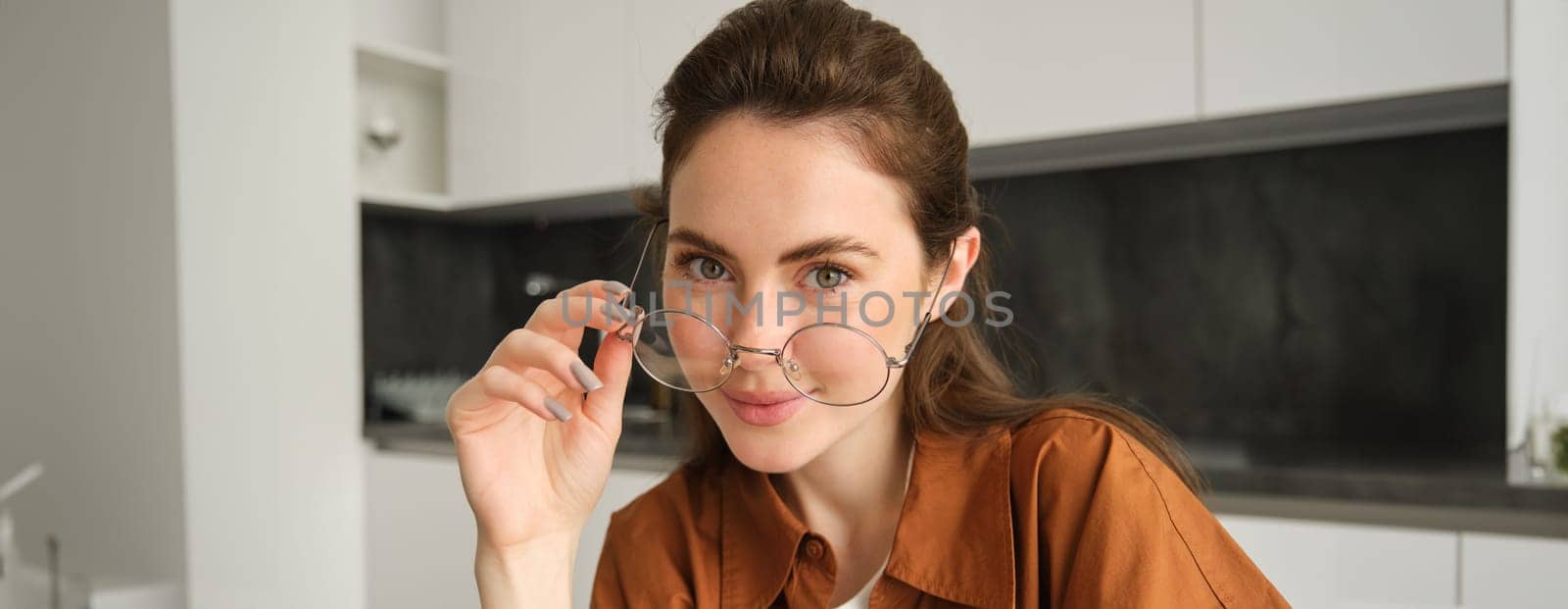 Portrait of beautiful young woman, smiling and looking happy, takes-off her glasses and looking at camera, sitting in kitchen at home by Benzoix