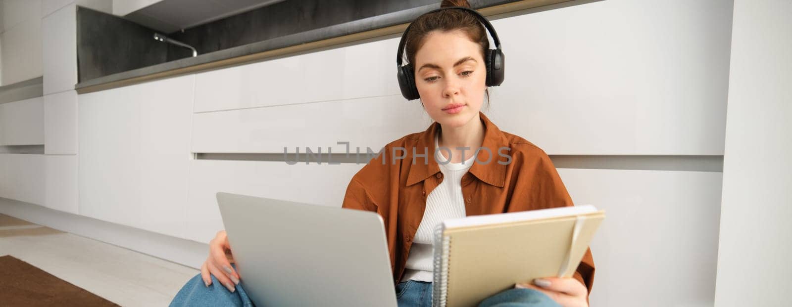 Portrait of young woman, modern student sits at home on floor and doing homework on laptop, listens music in headphones while making assignment or project for college.
