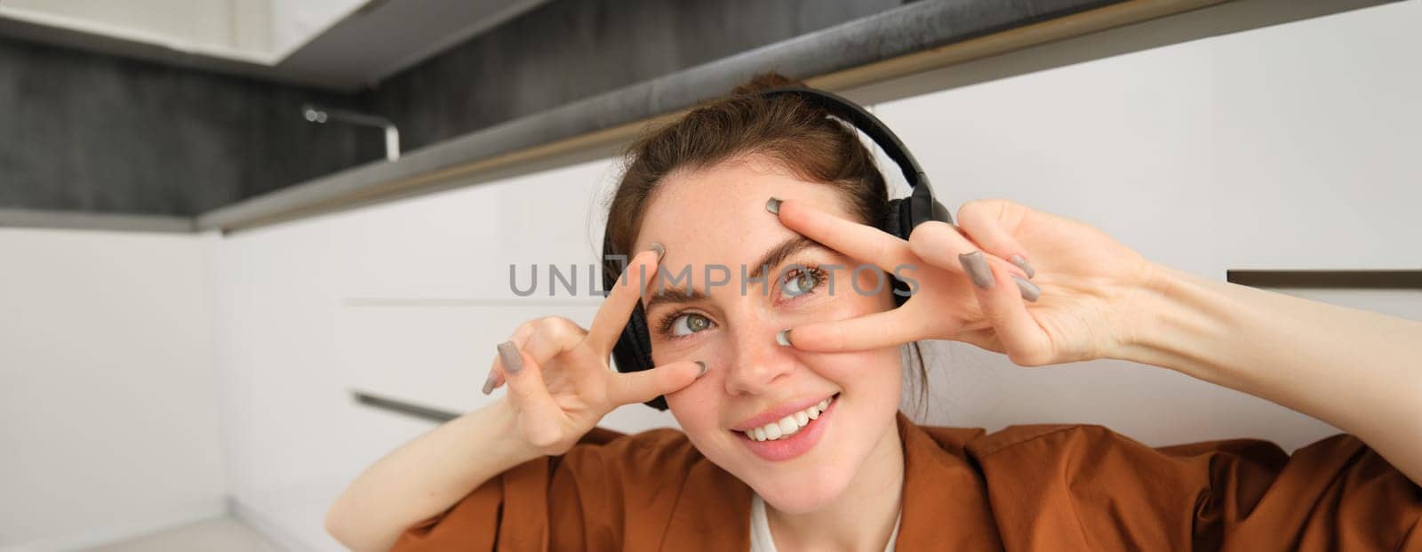 Close up portrait of woman in headphones, shows peace, v-signs near eyes, smiles and looks happy, sits on floor at home.