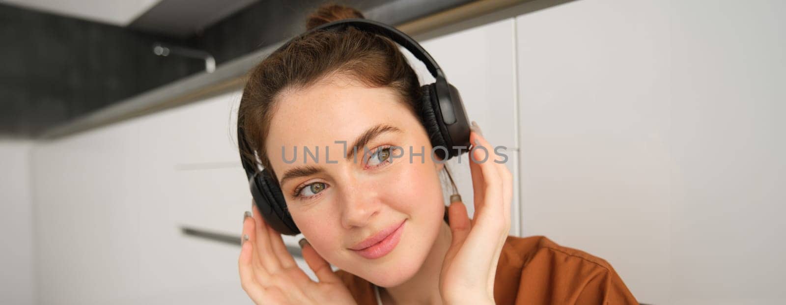Close up shot of cute young woman in wireless headphones, listens to music and smiles, relaxes at home with favourite songs playing on her playlist.