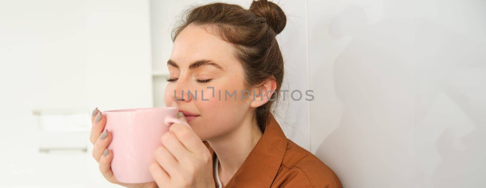 Close up portrait of beautiful brunette woman with cup of coffee, drinking aromatic beverage from mug, smiling from pleasure, sitting in kitchen at home.