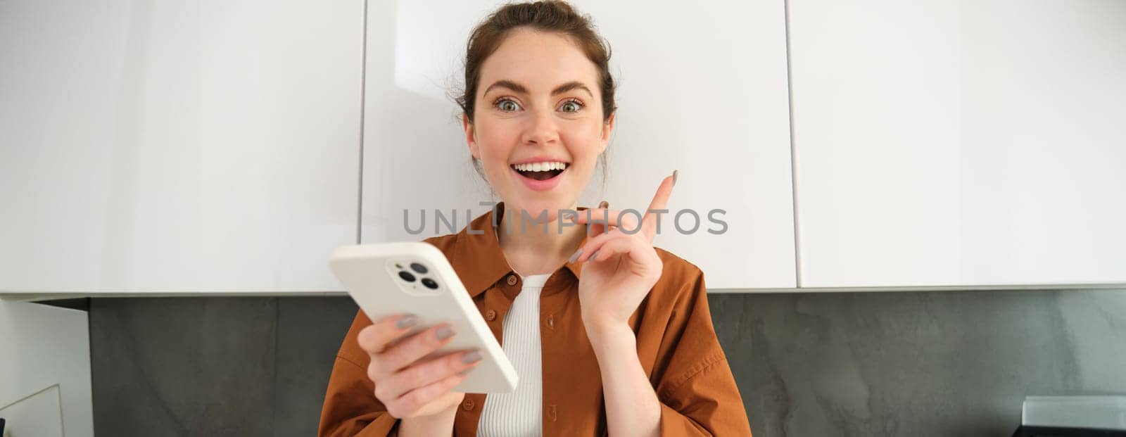 Portrait of excited young woman has an idea, holding smartphone and pointing finger up, standing in the kitchen at home by Benzoix