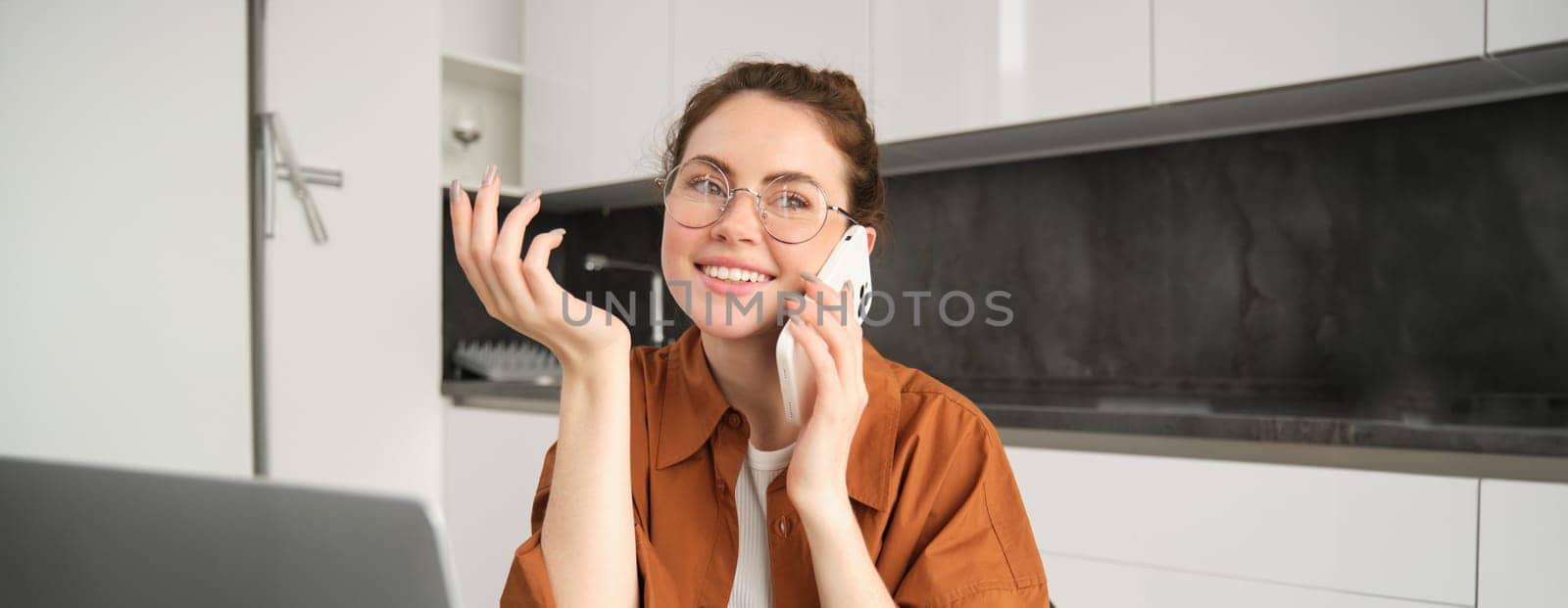 Portrait of young woman, business owner working from home, student making a phone call, sitting in kitchen with laptop, talking to someone.