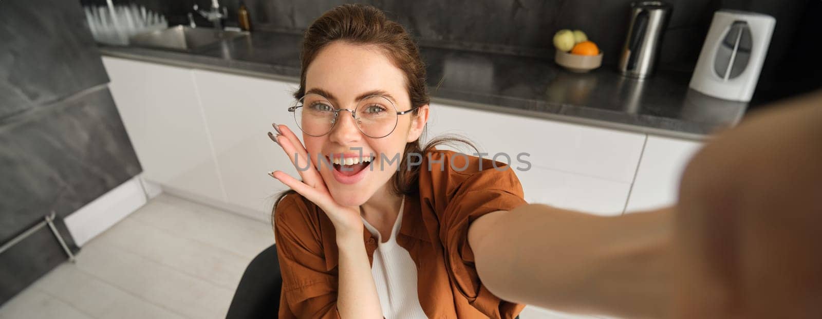 Portrait of carefree young woman in glasses, taking selfie from home, lifestyle blogger making photos in kitchen, laughing and smiling, using smartphone camera by Benzoix