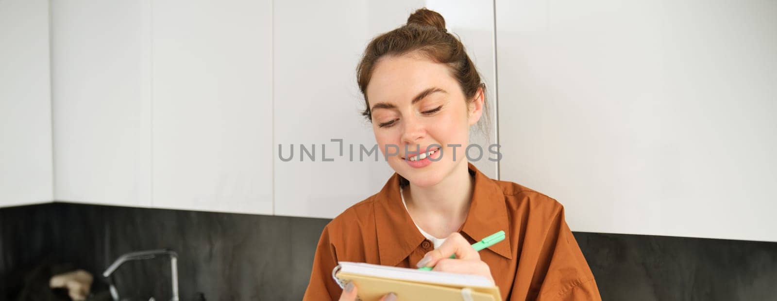 Portrait of young creative girl, writing down her ideas in planner, does homework in notebook, sits at home on kitchen counter.