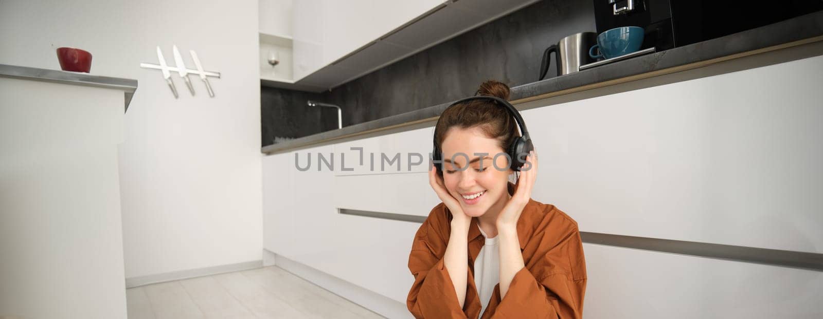Portrait of beautiful female model on kitchen floor at home, listens music in wireless headphones, smiling and looking happy.