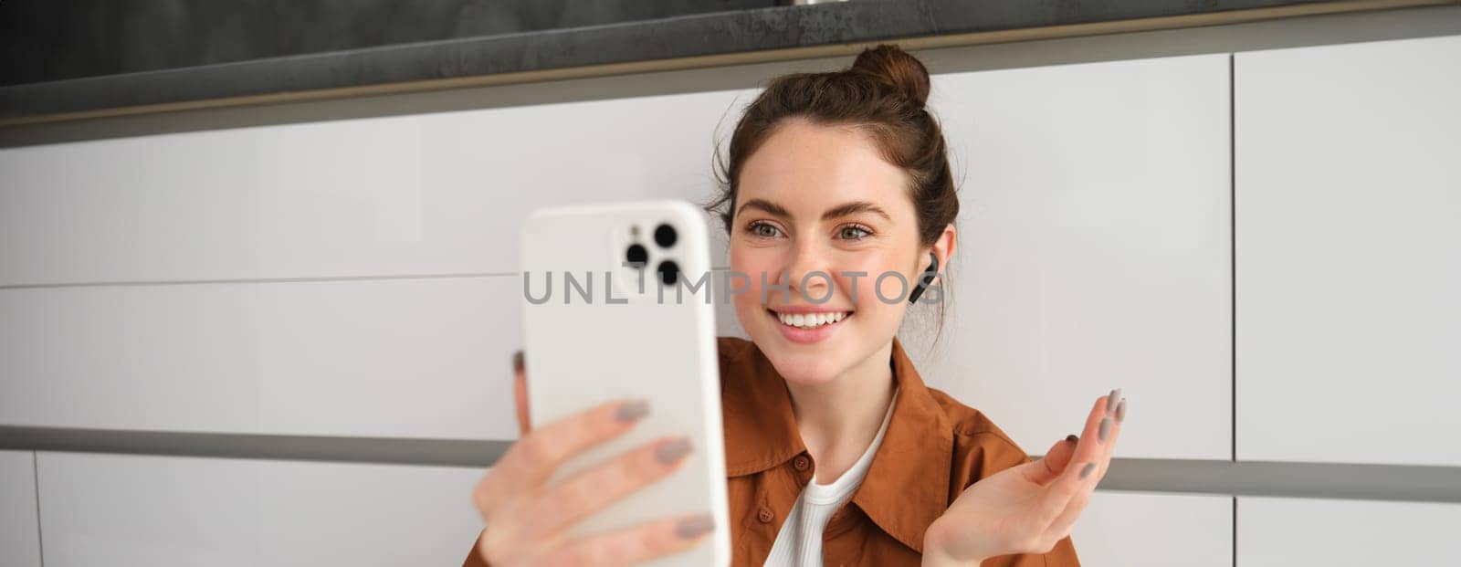 Portrait of beautiful smiling woman, talking to friend on smartphone video chat, connects to online conversation on mobile app, sitting on kitchen floor by Benzoix