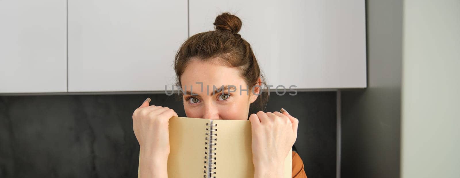 Lifestyle and people concept. Young brunette woman, holding notebook near face, hiding smile behind notepad and looking coquettish at camera by Benzoix