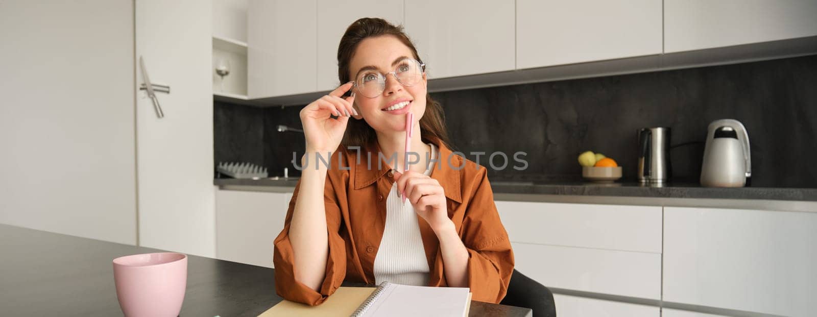 Portrait of creative young woman, writer making notes, teacher prepared papers for next lesson, sits at home in kitchen, looks up and thinks with pleased smile.