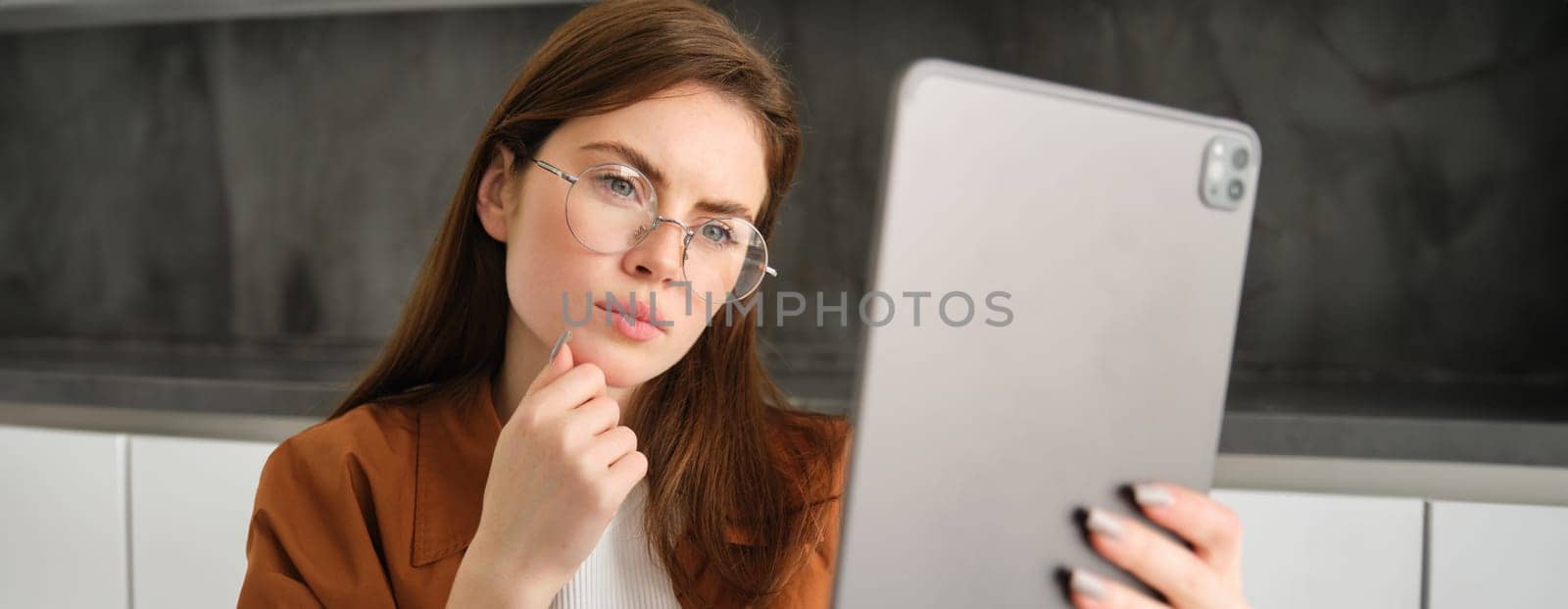 Portrait of working woman sits in kitchen, works from home, reads documents on digital tablet with thoughtful face , thinking, analysing papers.