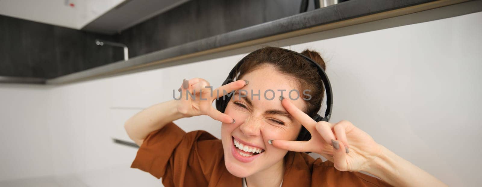 Carefree girl in wireless headphones, laughing and smiling, showing v-sign, peace gesture near face, listens to music, sits on floor at home.