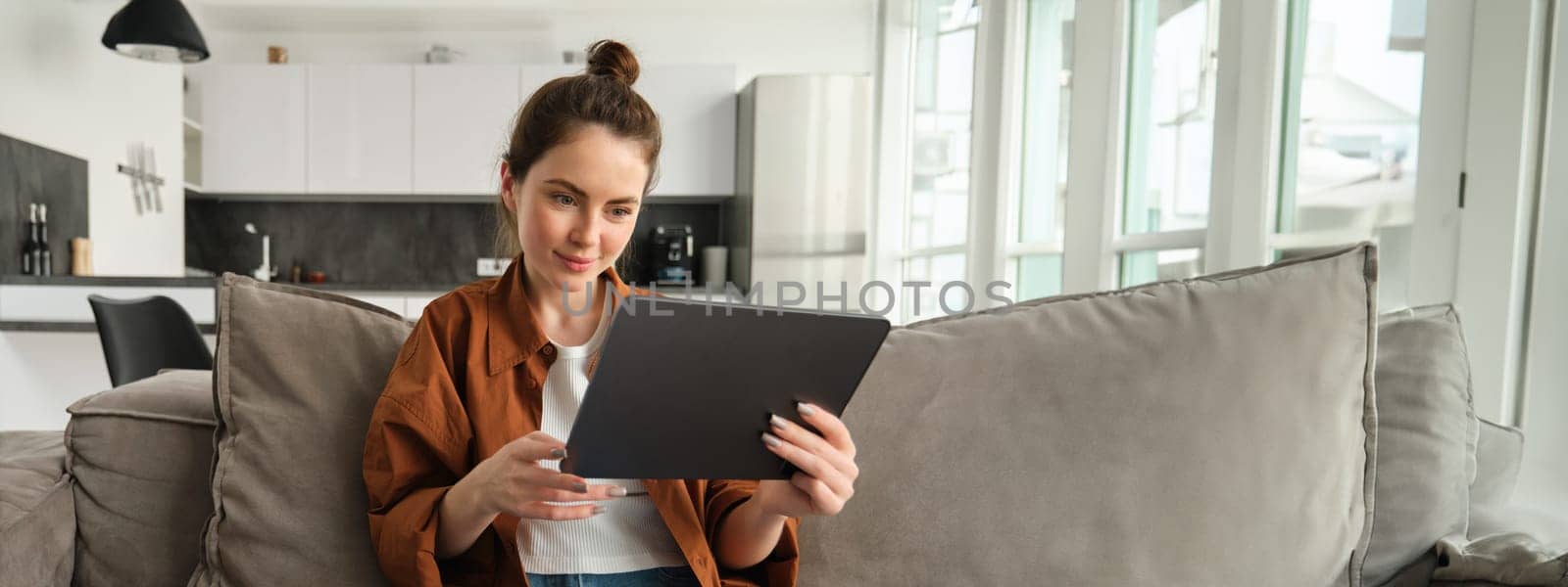 Portrait of smiling beautiful woman, sitting on couch, watching videos on digital tablet, reading e-book on her device, resting at home.