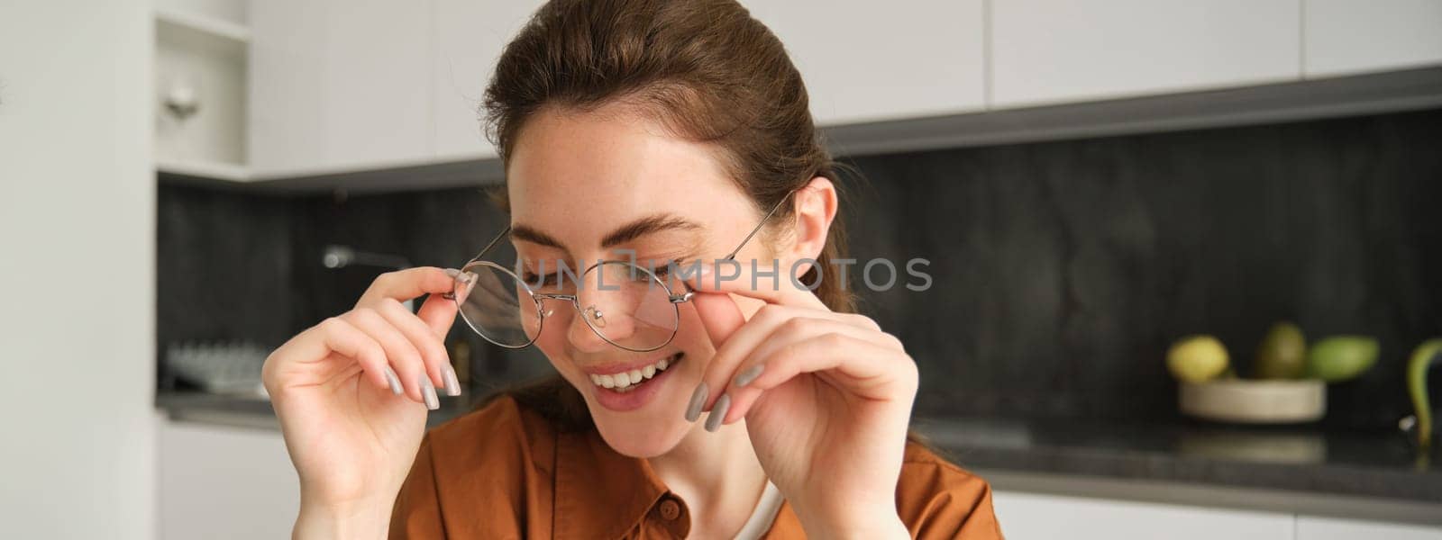 Portrait of young happy woman, sitting in kitchen, wearing glasses, reading or working from home by Benzoix