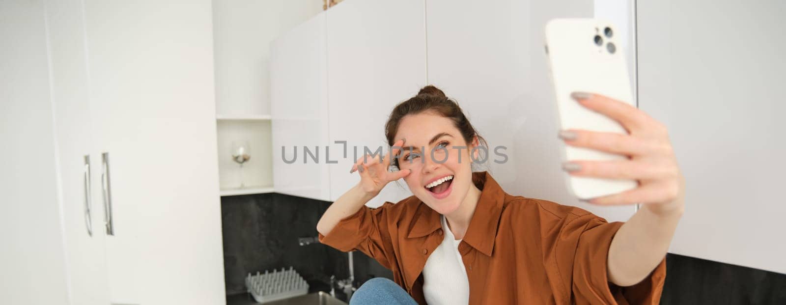 Young charismatic girl takes selfies in the kitchen, posing for photo, holds mobile phone with extended hand by Benzoix