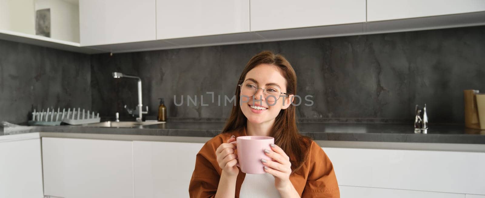 Cute smiling woman in glasses, sits in kitchen with cup of coffee, drinks tea from mug and looks happy, enjoys relaxing day at home.