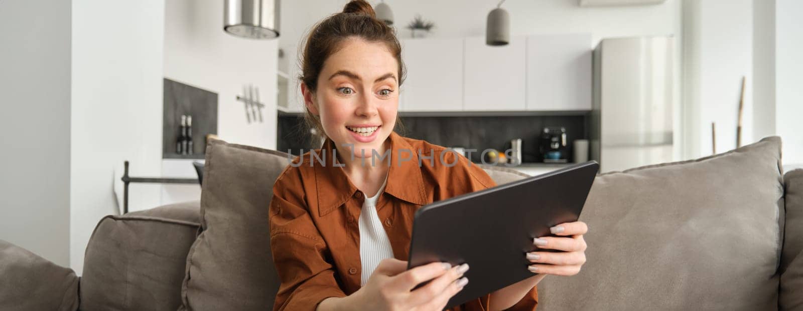 Portrait of excited, smiling young woman playing games on her digital tablet, tilting gadget and looking amused at screen, sitting on couch at home.