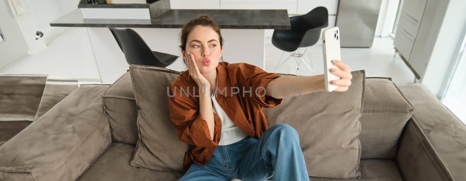 Portrait of cute smiling female model, takes selfie on smartphone app, posting photos on social media, using mobile phone camera, posing on sofa at home.