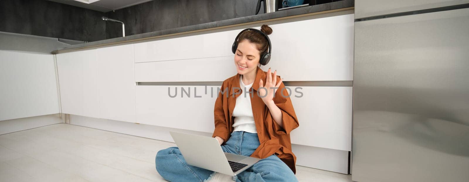 Portrait of happy woman in headphones, looking at laptop, waving hand at computer, chatting on video online, joins team meeting, calling friend on computer, sitting on floor.