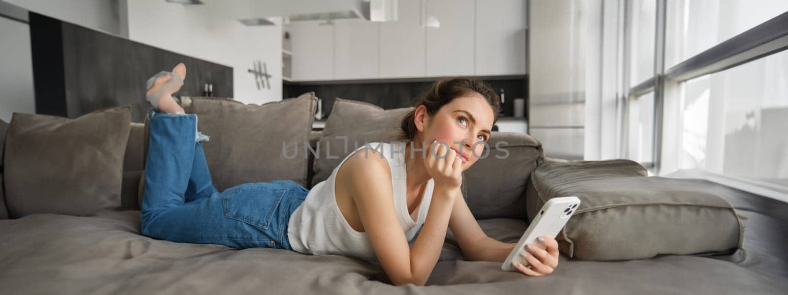 Online shopping concept. Young woman resting on couch, lying on sofa with smartphone, looking up and thinking.