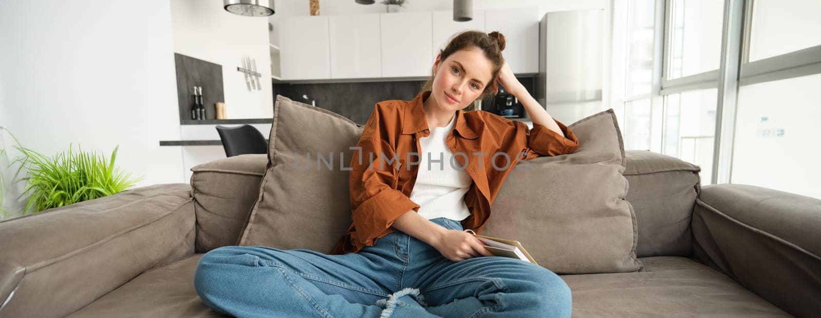 Portrait of young modern woman, female student sitting at home on sofa and looking at camera, posing on couch, smiling.
