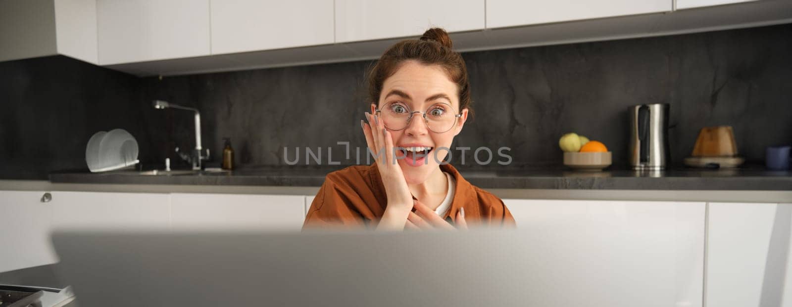 Close up portrait of amazed young woman, looking at laptop with happy, surprised face expression, sitting at home.