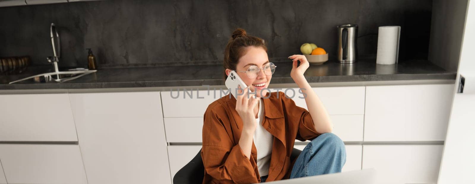 Excited young woman talking on mobile phone in front of laptop, sitting in kitchen with happy face expression, having a conversation.