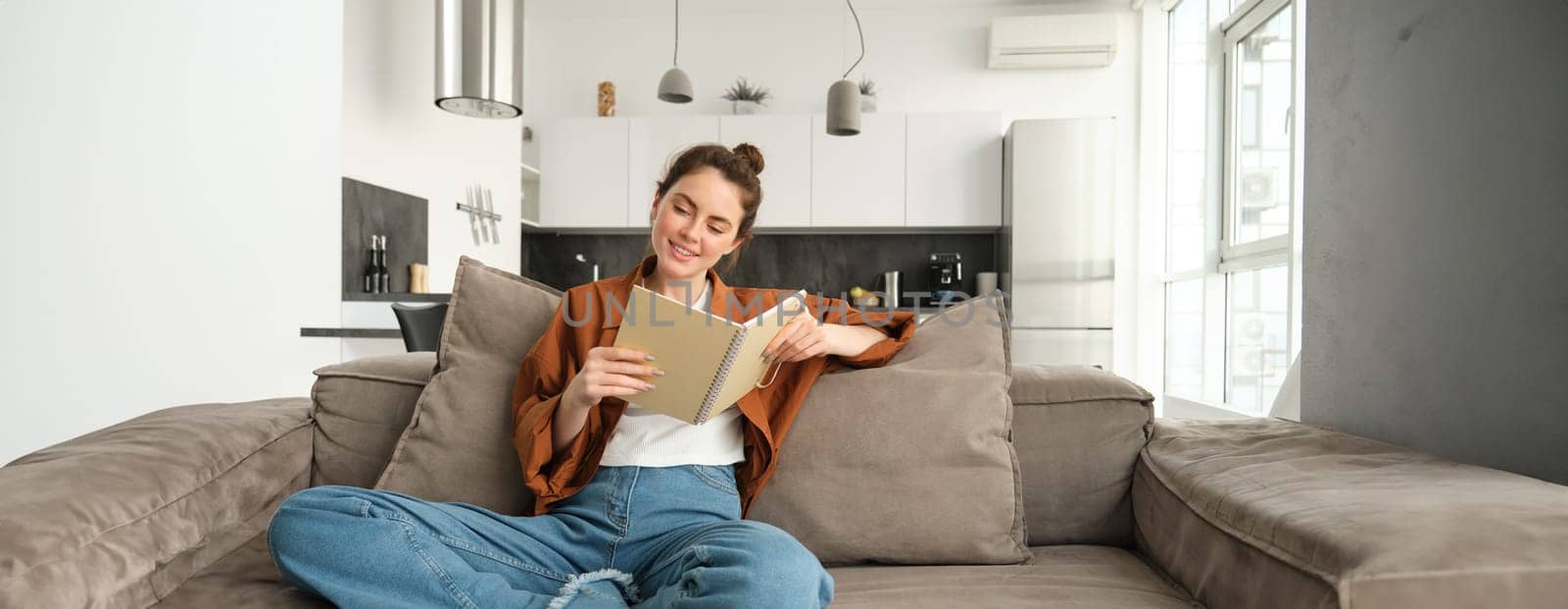 Beautiful young female student, sitting at home on sofa, relaxing on couch, reading notebook and smiling.